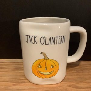 Image of a white mug resting on a table with "Jack O'Lantern" in black letters on the top of the mug and a orange pumpkin below the wording.